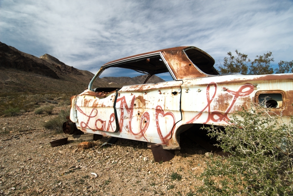 Death Valley, California photograph. An abandoned, graffiti'd car in Rhyolite, Nevada. It says 'Love Me' in red paint. Rhyolite was an old ore mining town near Death Valley that hit its heyday over a century ago. The buildings have crumbled, and now it's nothing but a beautiful ghost town. The car depicted is from a later era than the mining town, but still fits with the ghost town aesthetic. I highly encourage you to make Rhyolite part of your trip to Death Valley. 